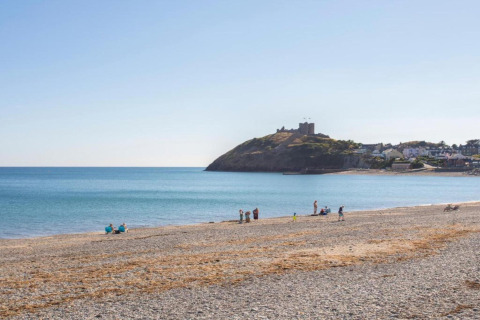 Plage de galets avec quelques personnes, mer bleue et château sur la colline près du site de glamping.