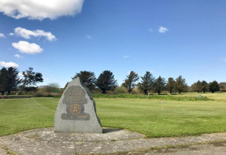 Monument en pierre sur pelouse verte d’un site de glamping, avec arbres et ciel bleu en arrière-plan.