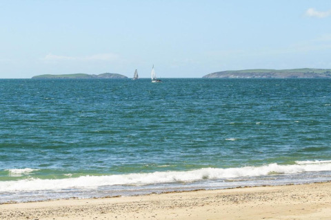 Sandy beach with gentle waves, two sailboats in blue sea, and distant green islands under a clear sky.