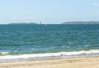 Sandy beach with gentle waves, two sailboats in blue sea, and distant green islands under a clear sky.