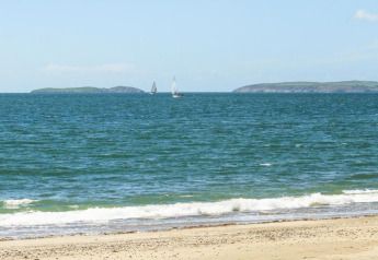 Blick auf einen Sandstrand, Wellen im Meer und zwei Segelboote, im Hintergrund grüne Inseln und Himmel.