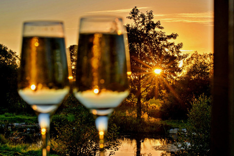 Deux verres de vin pétillant devant un coucher de soleil au bord d’un lac à la roulotte 'Elen' Gypsy Hut au Royaume-Uni.