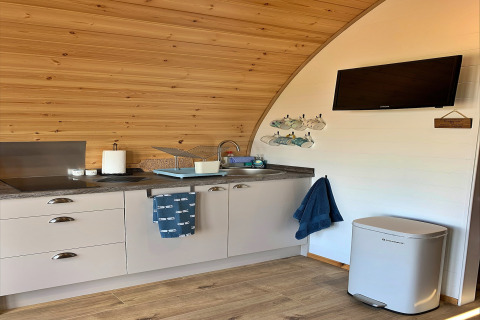 Modern kitchenette inside a wooden pod named 'Deep Dale' with TV, bin, towels, cups, and utensils.