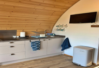 Modern kitchenette inside a wooden pod named 'Deep Dale' with TV, bin, towels, cups, and utensils.