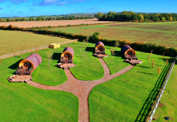 Luchtbeeld van vier glamping-pods op een groen terrein met wandelpaden en uitzicht op landelijke velden.
