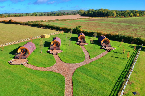 Aerial view of four modern glamping pods on a green field with paths and scenic countryside in the background.