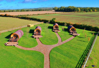 Aerial view of four modern glamping pods on a green field with paths and scenic countryside in the background.