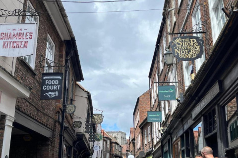 A vibrant street in the UK lined with historic brick buildings and shop signs like The Shop That Must Not Be Named.
