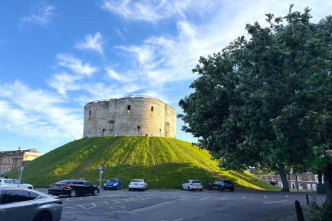 Round stone tower on green mound, surrounded by parked cars and trees under a blue, clouded sky.