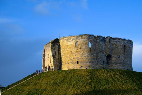 Round stone castle keep sits atop a grassy hill against a bright blue sky, historical building in view.