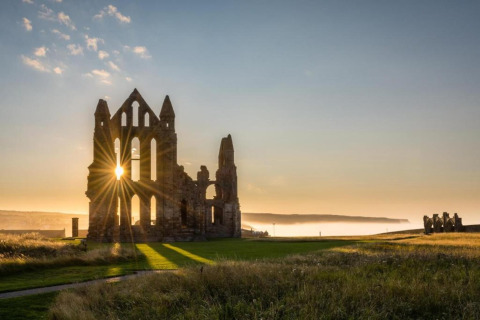 Sunrise shining through ancient ruins in a grassy field, under a clear sky with a few scattered clouds.