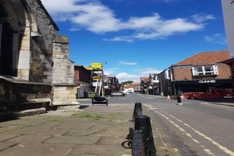 A street scene with historic stone buildings, shops, a ladder, and clear blue sky with white clouds.