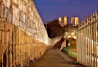 Vista serale di un sentiero illuminato lungo un muro cittadino e una cattedrale illuminata sullo sfondo.