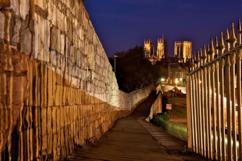 Evening view of a lit pathway along a city wall with a brightly illuminated cathedral in the distance.