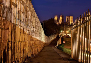 Evening view of a lit pathway along a city wall with a brightly illuminated cathedral in the distance.
