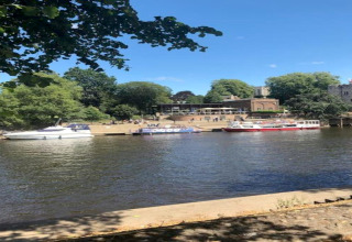 Barche ormeggiate lungo una riva tranquilla con alberi e edifici sullo sfondo in una giornata di sole.