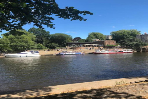 Boats docked along a calm riverbank on a sunny day, with trees and buildings in the scenic background.
