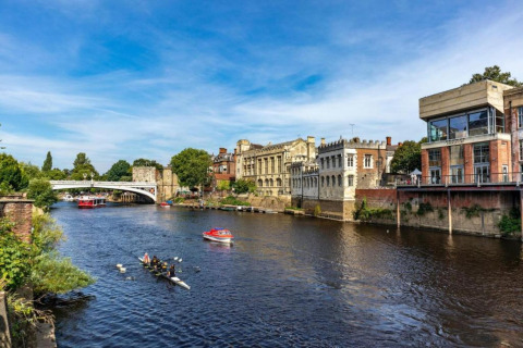 View of a river with rowers, boats, and historic riverside buildings under a clear blue sky.