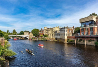 View of a river with rowers, boats, and historic riverside buildings under a clear blue sky.