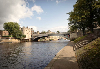 Rivierlandschap met een oude stenen brug en historische gebouwen, gezien vanaf het voetpad langs het water.
