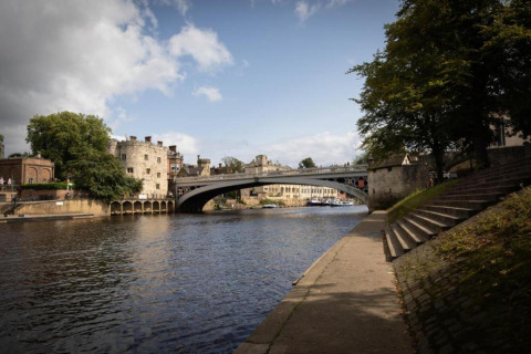 River scene with a historic stone bridge, old buildings in the background, and path along the riverside.