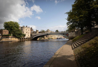 River scene with a historic stone bridge, old buildings in the background, and path along the riverside.