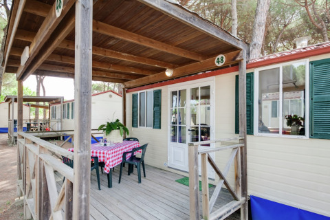 Stacaravan Luna Pineta in Italië met een overdekt houten terras, tafel, stoelen en pijnbomen.