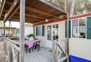 Stacaravan Luna Pineta in Italië met een overdekt houten terras, tafel, stoelen en pijnbomen.