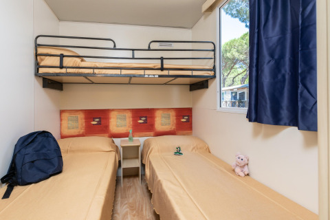 Kamer in Luna Pineta stacaravan in Italië met stapelbed, twee eenpersoonsbedden, raam en blauwe gordijnen.