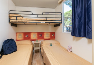 Kamer in Luna Pineta stacaravan in Italië met stapelbed, twee eenpersoonsbedden, raam en blauwe gordijnen.
