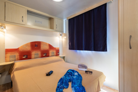 Slaapkamer in de Luna Pineta stacaravan in Italië met tweepersoonsbed, donkere gordijnen en kastruimte.