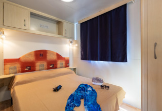Slaapkamer in de Luna Pineta stacaravan in Italië met tweepersoonsbed, donkere gordijnen en kastruimte.