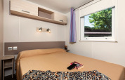 Slaapkamer in mobilhome Cottage «Cesenatico» in Italië met tweepersoonsbed, raam en kastjes.