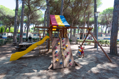 Speeltuin met glijbaan, schommels en klimwand bij Luna Pineta mobilhome in Italië, omringd door pijnbomen.