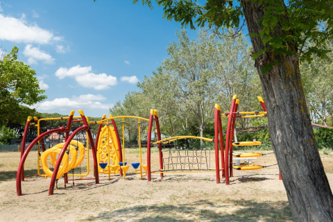 Spielplatz mit gelb-roten Geräten am Mobilheim Cottage «Cesenatico» in Italien, umgeben von Bäumen und blauem Himmel.