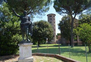 Standbeeld en historische toren bij Luna Pineta mobilhome, Italië, omgeven door groene bomen en zon.