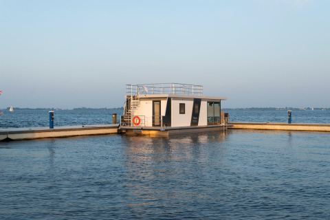 Casa flotante moderna llamada Houseboat Agua Dolce atracada en un puerto tranquilo, con agua y cielo al fondo.