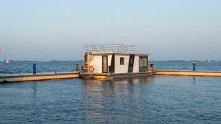 Modernes Hausboot namens Houseboat Agua Dolce vertäut in einem ruhigen Hafen, Wasser und Himmel im Hintergrund.