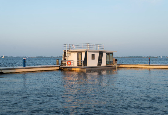 Casa flotante moderna llamada Houseboat Agua Dolce atracada en un puerto tranquilo, con agua y cielo al fondo.