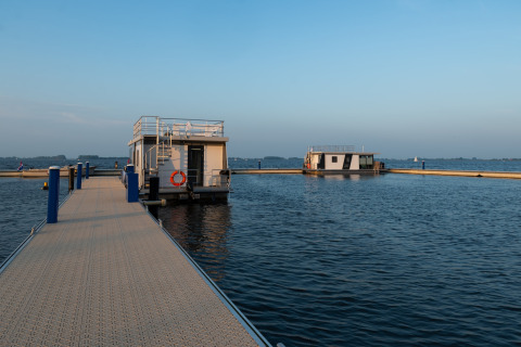 Foto del barco casa 'Agua Dolce' atracado en un muelle en Países Bajos, rodeado de agua tranquila y cielo azul.
