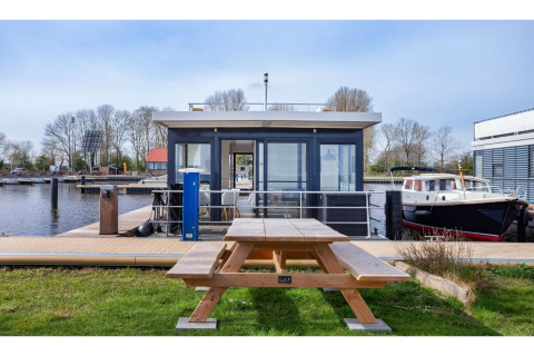 Casa flotante 'Agua Dolce' en Países Bajos con mesa de pícnic de madera y barcos al fondo del canal.