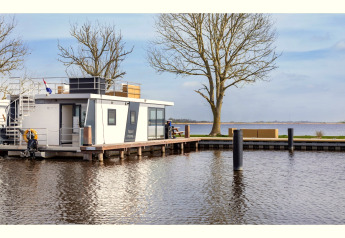 Hausboot ‘Agua Dolce’ in den Niederlanden, am Ufer vertäut mit Bäumen und Blick auf das Wasser.