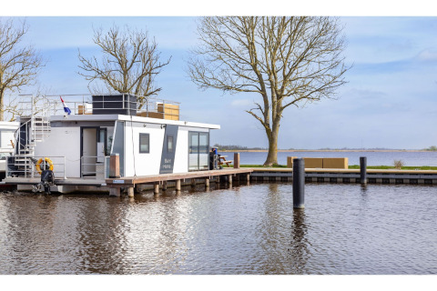 Hausboot ‘Agua Dolce’ in den Niederlanden, am Ufer vertäut mit Bäumen und Blick auf das Wasser.