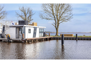 Hausboot ‘Agua Dolce’ in den Niederlanden, am Ufer vertäut mit Bäumen und Blick auf das Wasser.