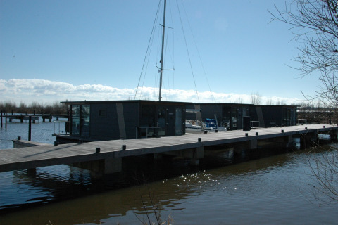 Modern houseboat at a dock in the Netherlands, surrounded by water, with a blue sky and sailing mast.