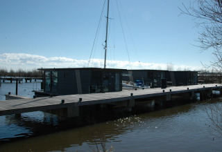 Modern houseboat at a dock in the Netherlands, surrounded by water, with a blue sky and sailing mast.