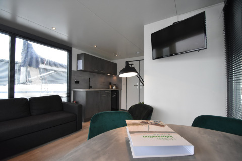 Modern houseboat interior in the Netherlands featuring a kitchen, sofa, TV on the wall, and large windows.