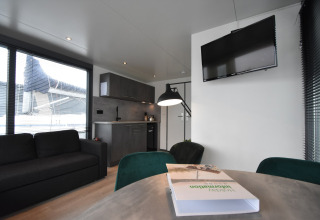 Modern houseboat interior in the Netherlands featuring a kitchen, sofa, TV on the wall, and large windows.
