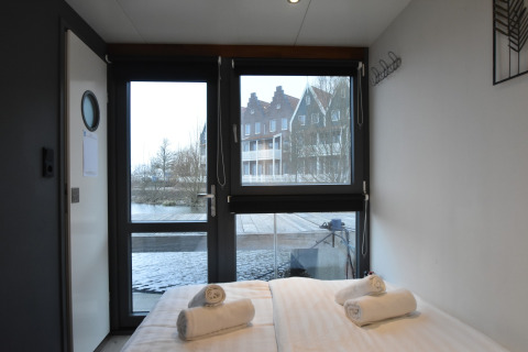 Bedroom on houseboat 'Boat' in the Netherlands with a water and houses view through large windows.
