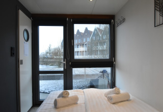 Bedroom on houseboat 'Boat' in the Netherlands with a water and houses view through large windows.